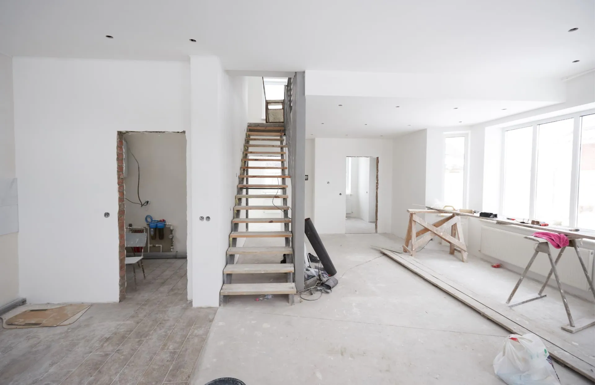 Interior of a home mid-renovation, drywalled walls and a newly built staircase with the subfloor still exposed and trestles set up for work.